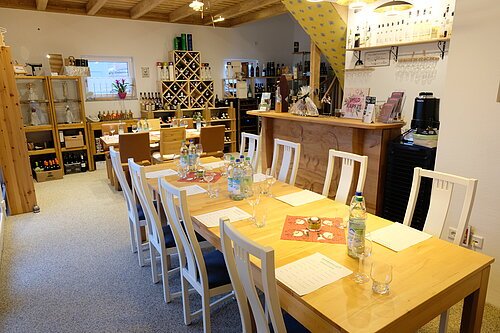 Gedeckter Tisch für eine Weinverkostung in der Weinhandlung Barbaricum. Acht weiße Stühle um einen großen Holztisch in einem hellen Raum. Im Hintergund Holzregale mit Wein und anderen Flaschen.